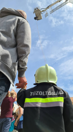 Felnőtt és tűzoltóruhás, sisakos gyermek felfelé néz a magasbó mentőre, alsó gépállásból