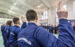 A kép előterében eskühöz emelt kézzel egy hallgató, a válla felett az eskü szövegét előolvasó hallgató